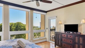 A bedroom with a large window, a ceiling fan, a bed with a floral bedspread, a dresser with a flat-screen TV, and a balcony with a view of trees and buildings.