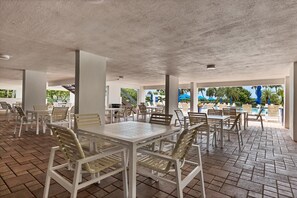 Un área de estar al aire libre con mesas y sillas, una piscina al fondo y un paseo cubierto.