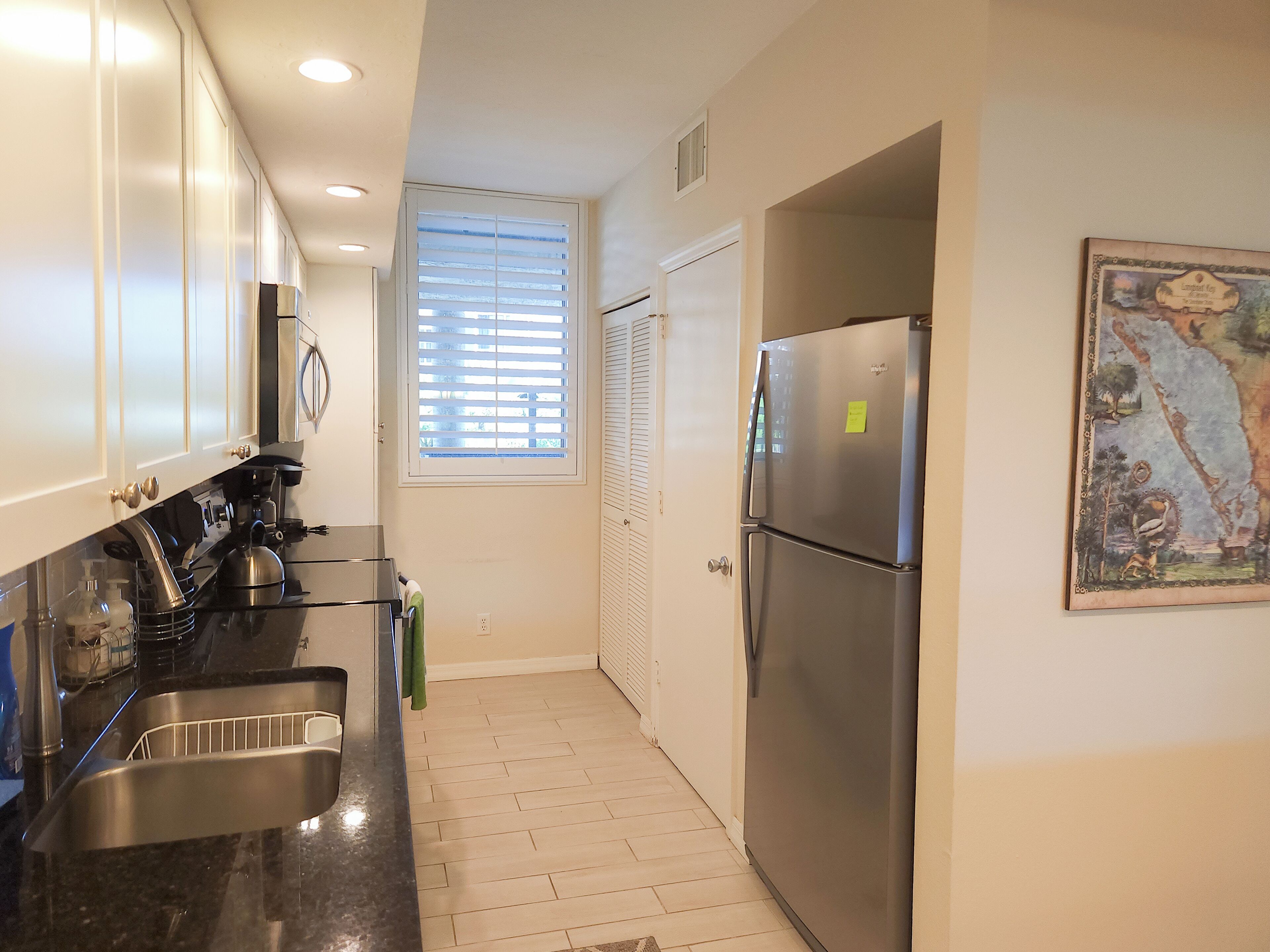 A modern kitchen with stainless steel appliances, a window with blinds, and a painting on the wall.