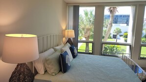 A hotel room with a bed, two bedside lamps, and a view of palm trees and a building through large windows.