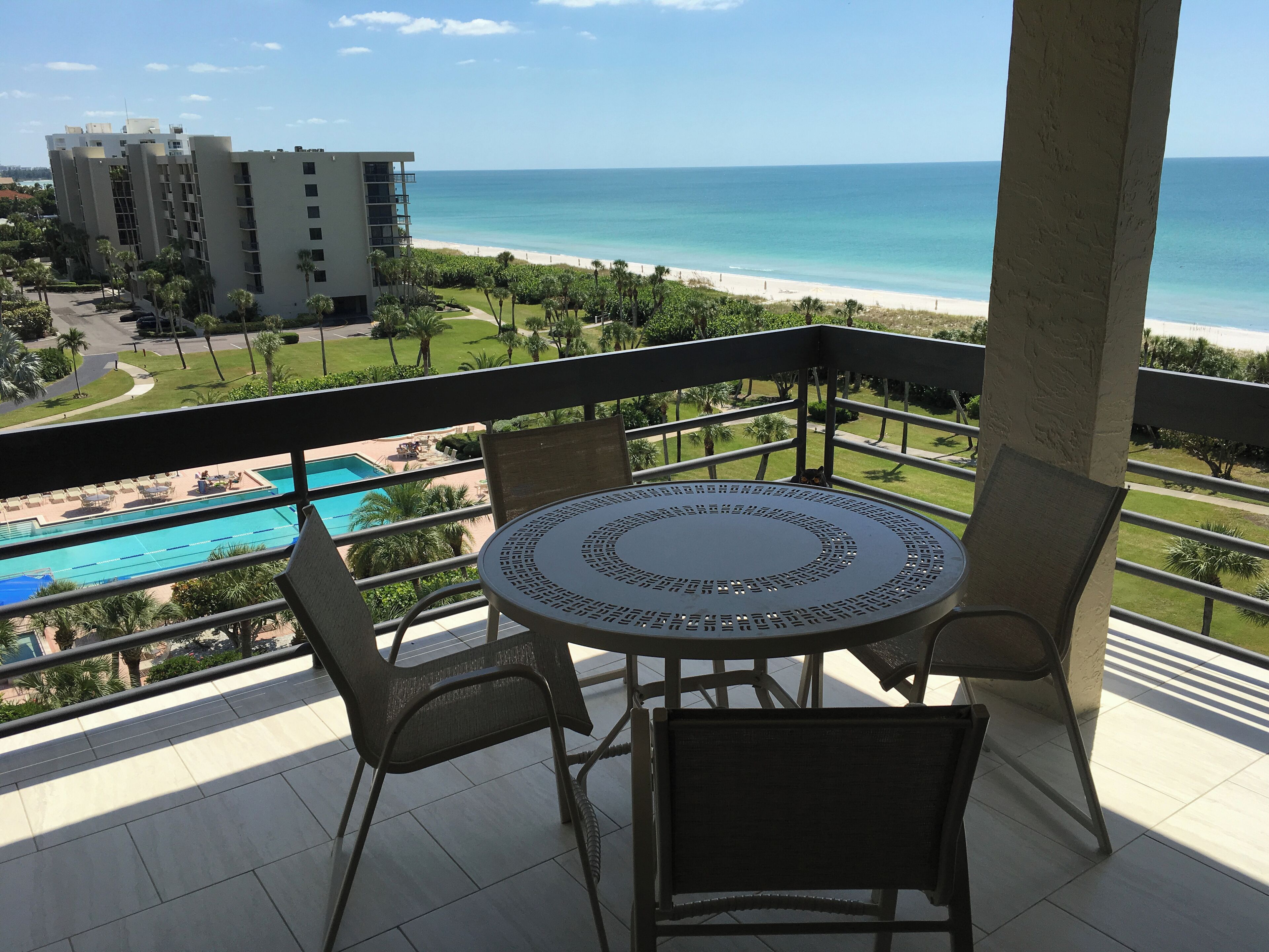 Ein Balkon mit rundem Tisch und Stühlen, der Blick auf einen Strand und ein Schwimmbecken freigibt.