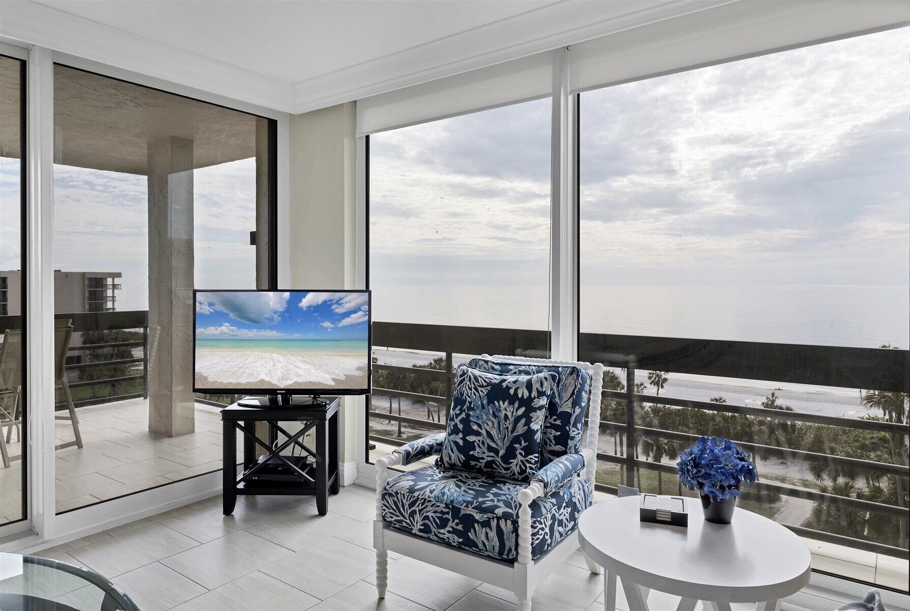 A balcony with a large TV, a patterned chair, and a small table with a vase.
