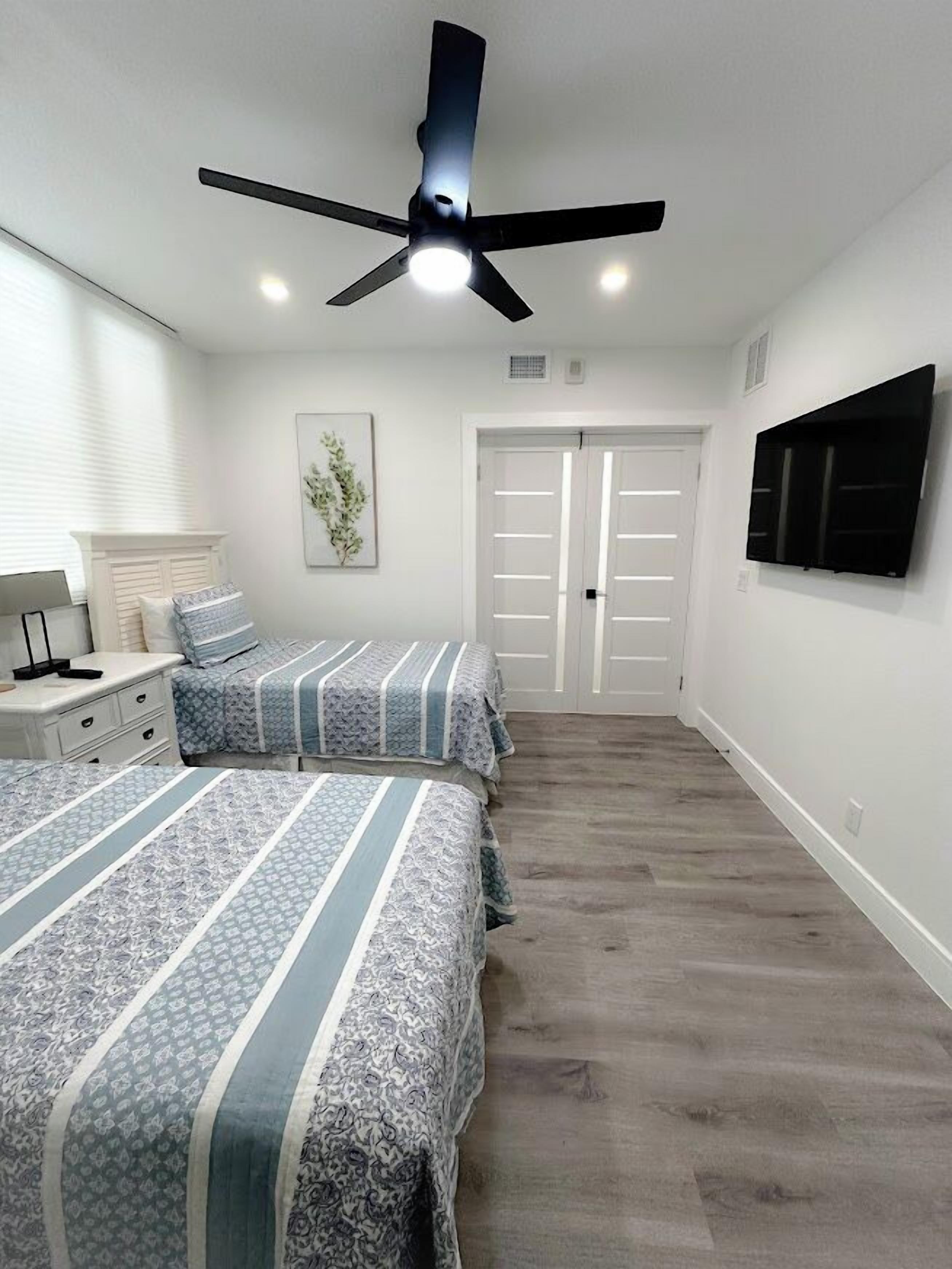 Un dormitorio con dos camas, un ventilador de techo, un televisor montado en la pared y una puerta que da a otra habitación.