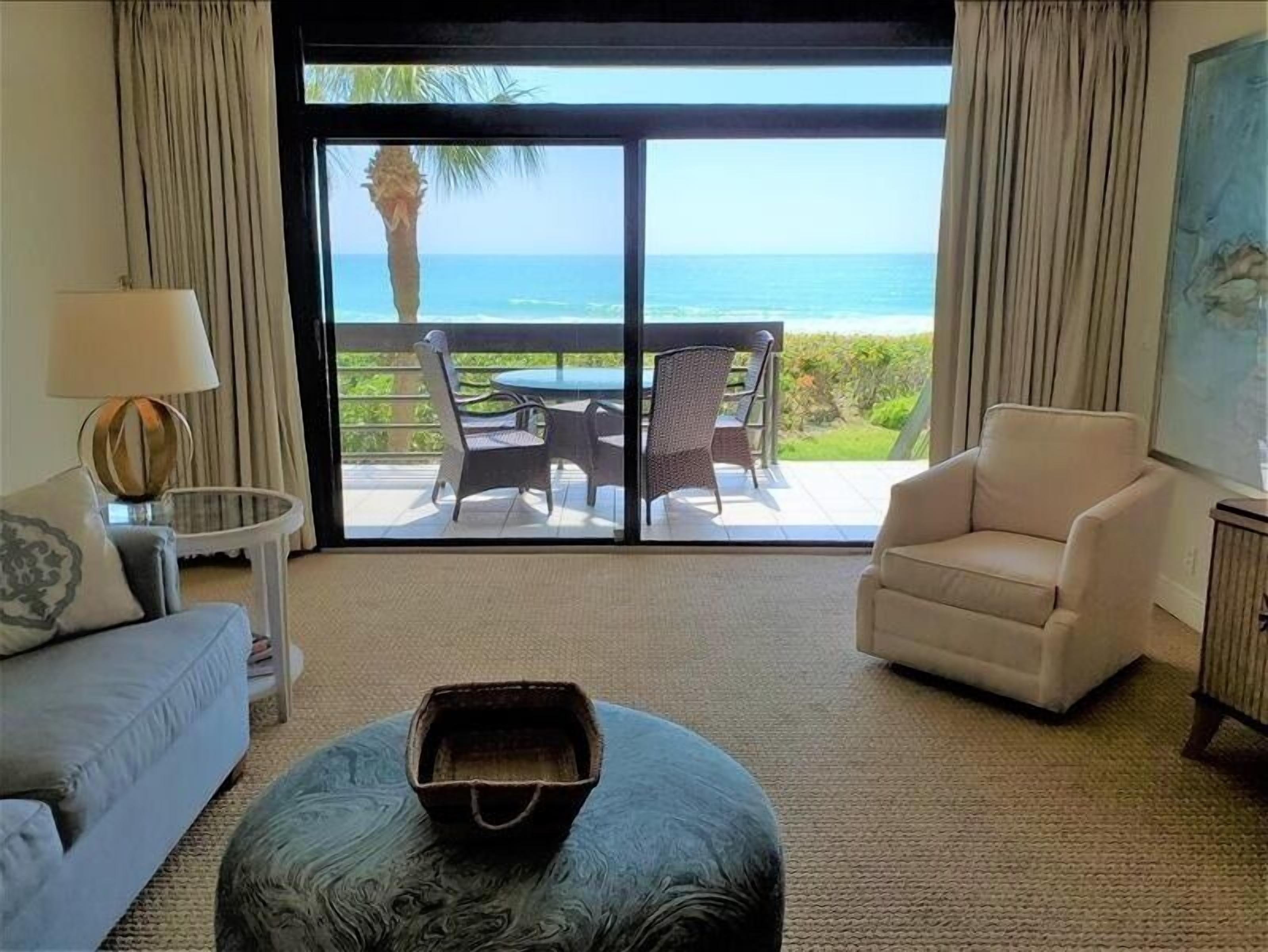 Quarto com vista para o oceano, uma poltrona branca, uma mesa redonda com cadeiras e um abajur.