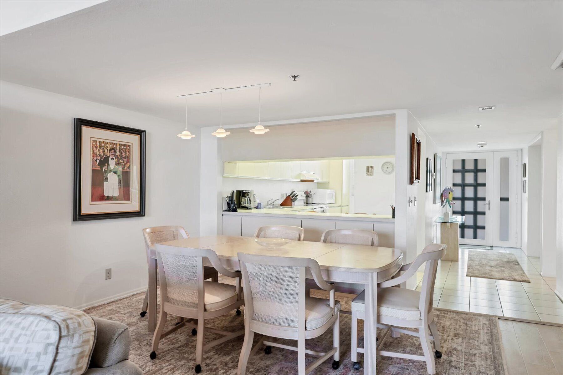 A modern dining area with a white table and chairs, a kitchen in the background, and a framed picture on the wall.