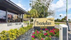 A beach-themed sign with the address 1045-1145 GMD, surrounded by landscaped gardens and a parking area. - 2/2 Condo BEACH VIEW, Pool, Fitness, Tennis (Longboat Key)