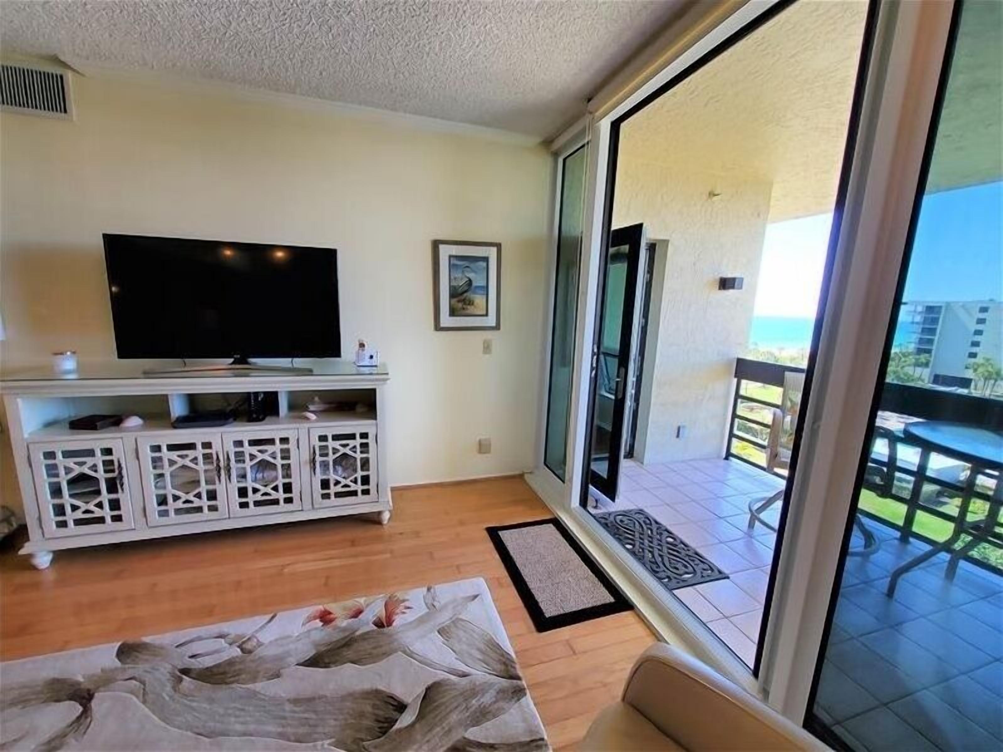 A living room with a flat-screen TV, a white entertainment unit, a framed picture, and a view of the balcony and outdoor area.