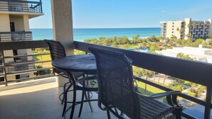 A balcony with two wicker chairs and a table, overlooking a beach and a golf course with palm trees.