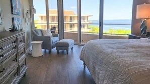 Un dormitorio con una cama grande, una silla, una mesita y un aparador. Hay ventanas amplias con vistas a un edificio y al océano.