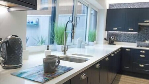 A modern kitchen with dark cabinets, a white countertop, a kettle, a coffee mug, and a window with a view of a building and greenery outside. - Jalebo (Cleethorpes)
