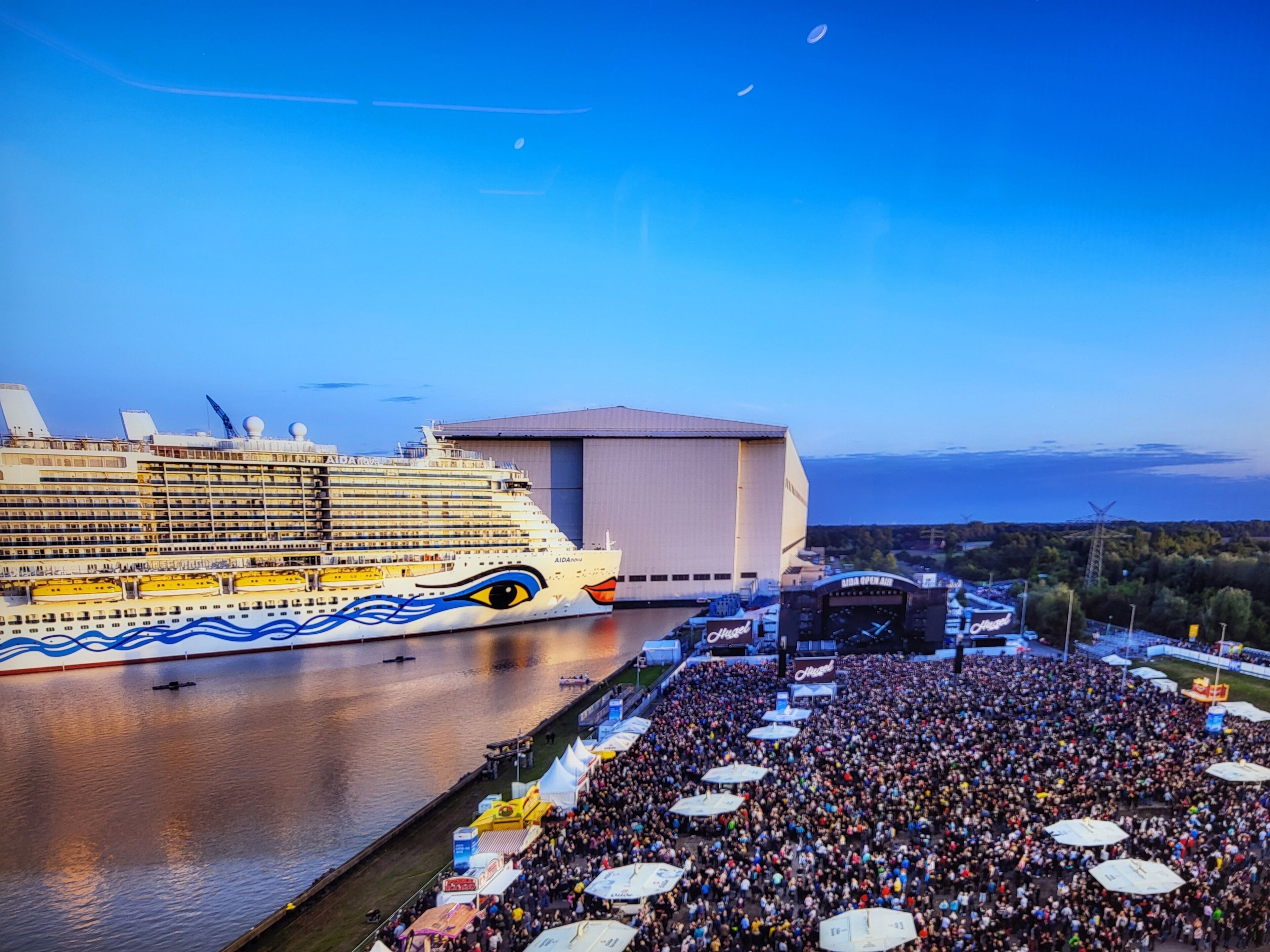 A cruise ship docked at a port with a large crowd gathered in the foreground.