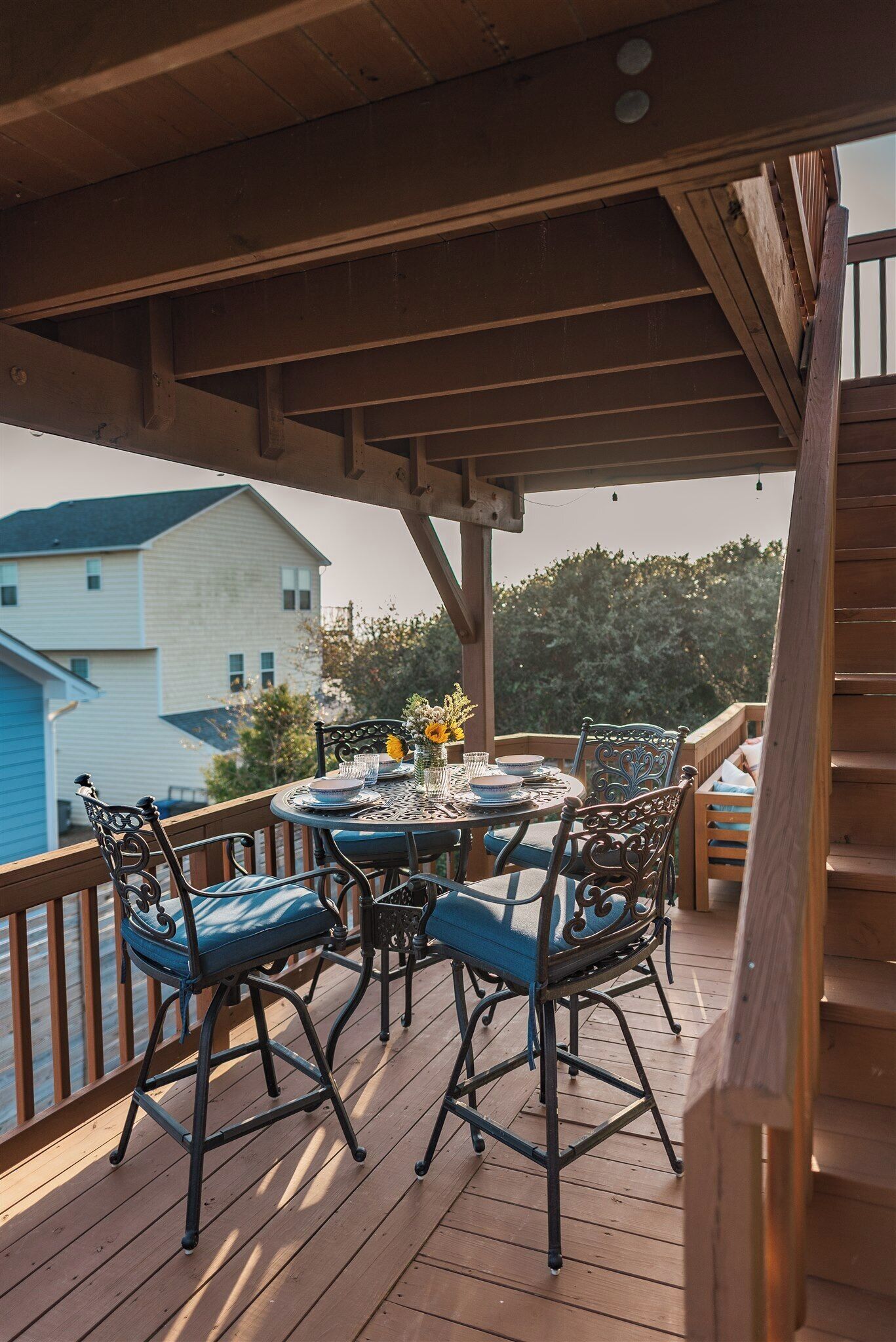 A wooden deck with a round table and chairs, a railing, and a view of a house in the background.