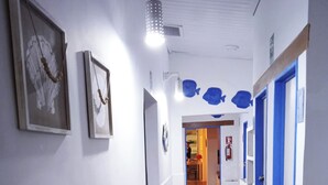 A hallway with framed artwork on the walls, a blue door on the right, and a wooden bench at the end. - Posada Lagunita by Mediterraneo (Los Roques)