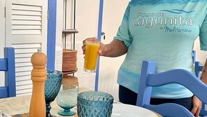 A person holding a glass of orange juice at a table with blue chairs, a white plate, and a blue glass. - Posada Lagunita by Mediterraneo (Los Roques)