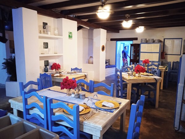 Un espace repas avec plusieurs tables et des chaises bleues, chaque table étant dressée avec des assiettes, des couverts et des fleurs rouges.