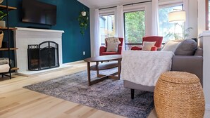 A living room with a fireplace, a red armchair, a grey sofa, and a large window with curtains. - Charming 4-bedroom family home in Beaverton with AC, fitness room, WiFi (Beaverton)