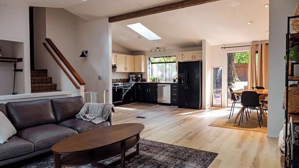 A spacious living area with a sofa, coffee table, and a dining table with chairs. A kitchen with modern appliances and cabinets is visible in the background. - Charming 4-bedroom family home in Beaverton with AC, fitness room, WiFi (Beaverton)
