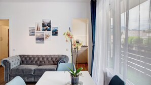 A modern living room with a grey tufted sofa, a white dining table, and a potted orchid. There are framed pictures on the wall and a large window with curtains. - Gemütliche und Ruhige Wohnung im Zentrum von Oberammergau! (Oberammergau)