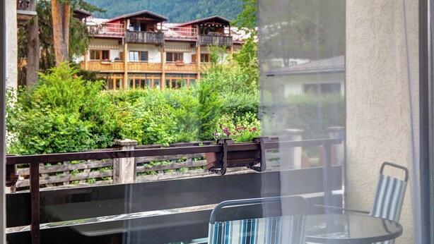 A balcony with a glass table and chairs, overlooking a wooden building with balconies and a lush green landscape. - Gemütliche und Ruhige Wohnung im Zentrum von Oberammergau! (Oberammergau)