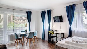 A modern hotel room with a wooden floor, a bed with white linens, a desk with a chair, a TV on the wall, and large windows with blue curtains. - Gemütliche und Ruhige Wohnung im Zentrum von Oberammergau! (Oberammergau)
