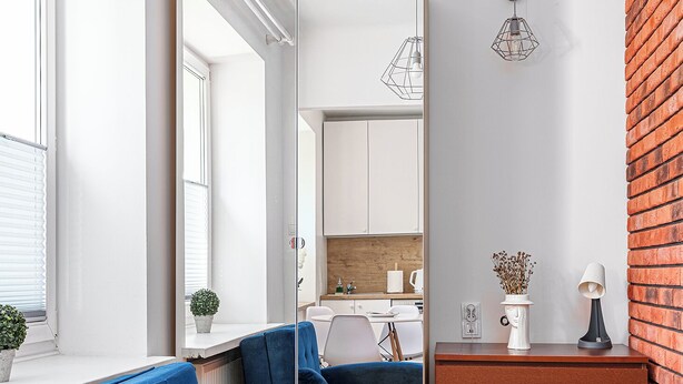 A modern living room with a blue sofa, a wooden floor, a brick accent wall, and a kitchen in the background. - RentPlanet - Apartament Obrzeżna (Warsaw)