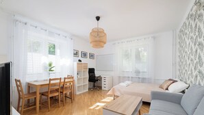 A modern, well-lit room with a wooden dining table, a grey sofa, a bed with a white and brown pillow, and a large window with sheer curtains. - RentPlanet - Apartament Obrzeżna (Warsaw)