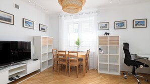 A modern living room with a TV, wooden dining table, chairs, and a black office chair. There are white shelves, a wicker lamp, and a window with curtains. - RentPlanet - Apartament Obrzeżna (Warsaw)