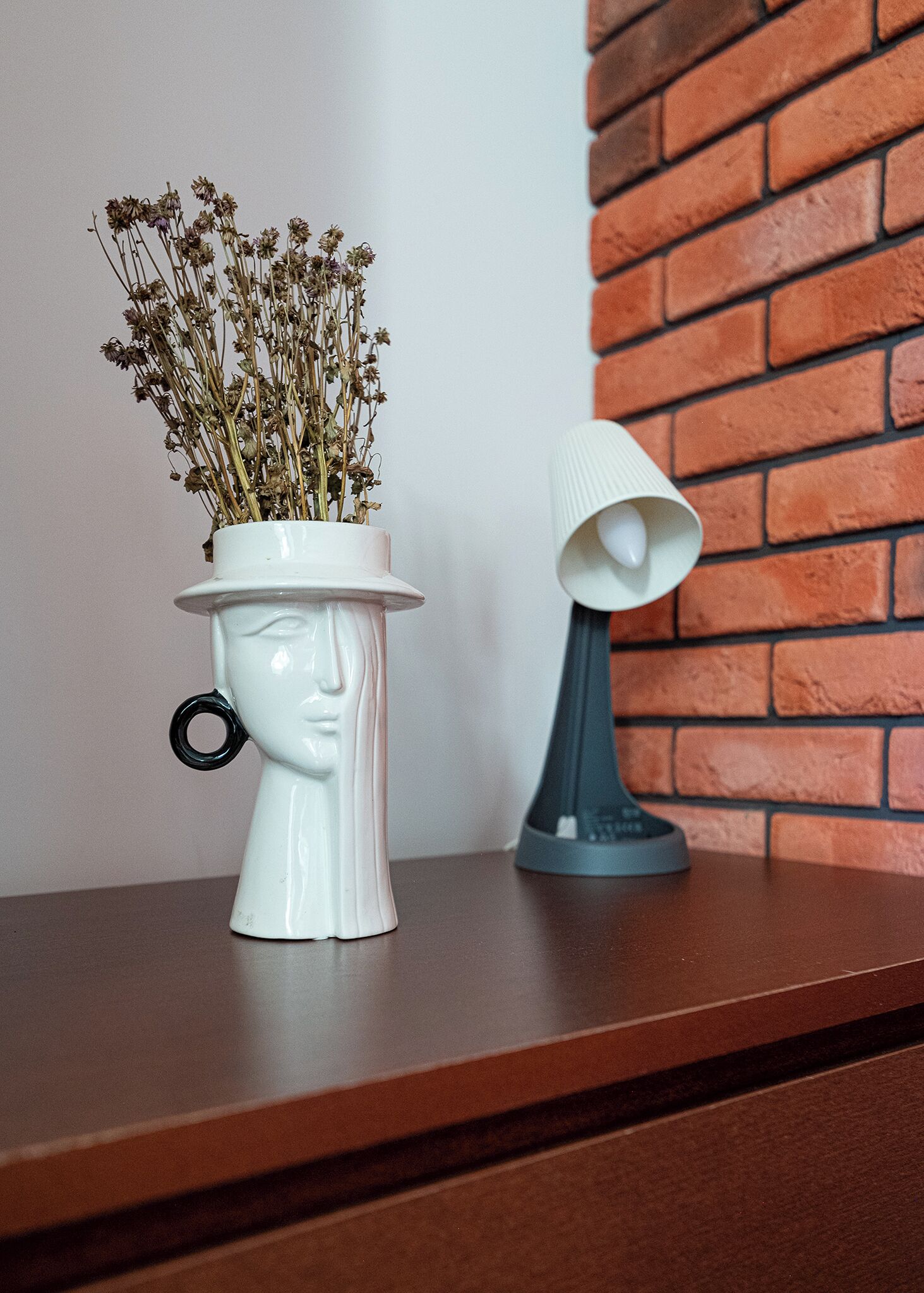 A white ceramic vase with dried flowers on a wooden shelf against a brick wall.