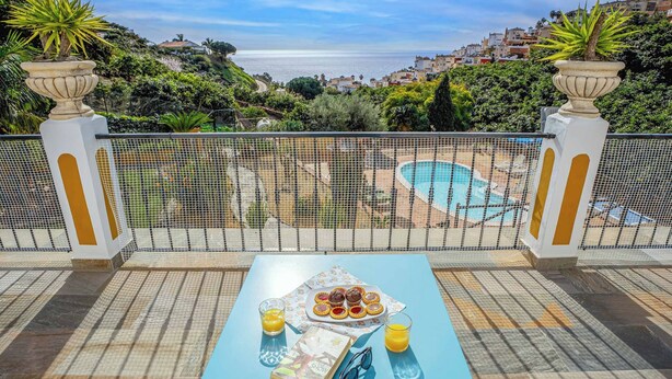 A terrace with a pool, a table with a book, glasses, and a plate of pastries, overlooking a coastal town.