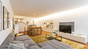 A modern living room with a grey sofa, a dining area with a wooden table and chairs, a staircase, and a flat-screen TV mounted on the wall.