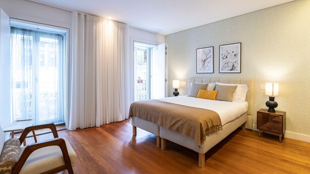 A hotel room with a large bed, two bedside lamps, a wooden nightstand, a chair, and a view of the outside through a window with sheer curtains.