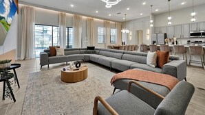 Una sala de estar moderna con un sofá seccional gris, una mesa de centro redonda de madera y un comedor con una cocina al fondo.