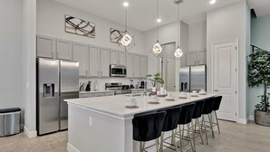 A modern kitchen with white cabinets, stainless steel appliances, a central island with bar stools, and pendant lighting. - Epic Paradiso Mansion-Pool/Game Room/Theater-2 (Orlando)