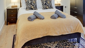 A neatly made bed with a beige bedspread, two pillows with a floral pattern, and two rolled towels. There is a dark-colored wardrobe to the right, a nightstand with a lamp, and a framed picture on the wall above the bed. - Harrow Bank Cottage Family and Pet Friendly Coastal Escape in Tweedmouth (Tweedmouth)