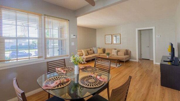 A spacious living room with a large sofa, a dining table with placemats, and a ceiling fan. - Centrally Located Cottage Near Lake Trails Shops (La Mesa)
