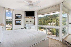 A hotel room with a large bed, a ceiling fan, a TV, and a view of a mountainous landscape.