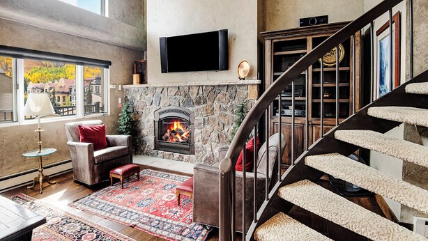 A spacious living room with a stone fireplace, a large window with a city view, and a modern staircase. - Vantage Point Condo 304 | Spacious 3BR in Lionshead Village (Vail)