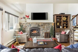 A living room with a stone fireplace, a flat-screen TV mounted above it, and a staircase leading to an upper level.