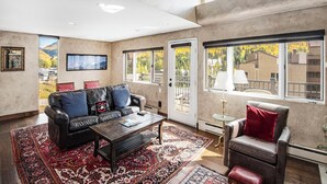 A spacious living room with a leather sofa, a coffee table, and a red patterned rug. There are large windows offering views of a mountainous landscape. - Vantage Point Condo 304 | Spacious 3BR in Lionshead Village (Vail)