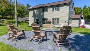 Een gebouw met twee verdiepingen, een grind terras, drie Adirondack stoelen en een kleine vuurplaats.