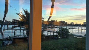 Vista a través de una ventana de una zona costera con palmeras, un muelle y un atardecer.