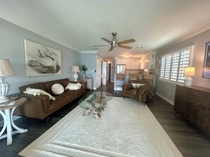 A spacious living room with a brown sofa, a glass coffee table, and a large rug. A kitchen is visible in the background with white cabinets and a refrigerator. - Ground level 2-bedroom condo with WiFi in wonderful location Panama City Beach (Panama City Beach)