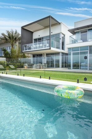 A modern two-story house with a swimming pool, a balcony with outdoor furniture, and a clear sky.