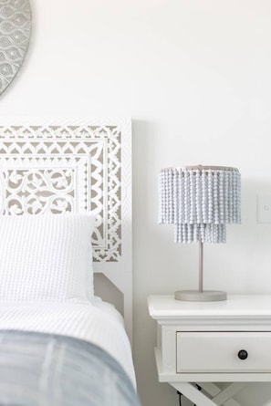 Ein weißes Bett mit einem gemusterten Kopfteil, ein Nachttisch mit einer Lampe und ein an der Wand befestigter Lichtschalter.