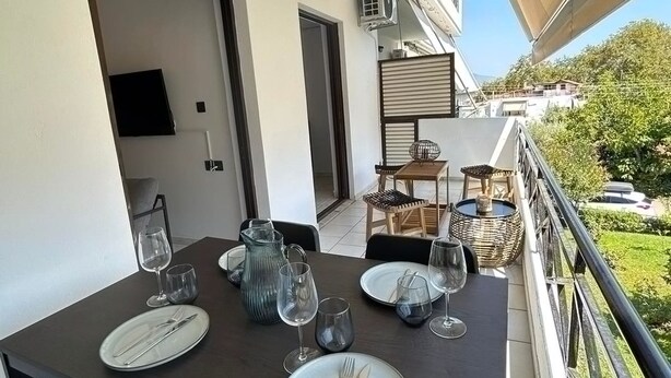 Une table à manger dressée avec des assiettes, des couverts et des verres, sur un balcon offrant une vue sur la verdure et les bâtiments.