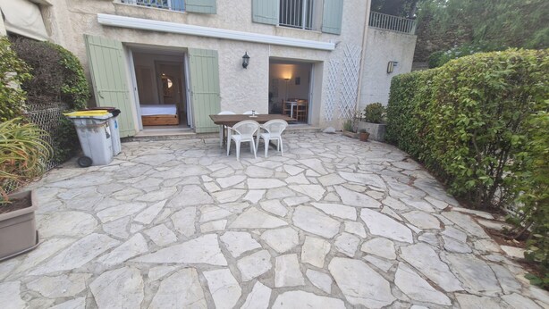 A stone-paved patio with a table and chairs, a trash bin, and a building with green shutters. - Résidence La Belle Poule 1172 (Sainte-Maxime)