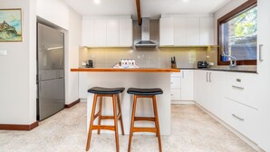 A modern kitchen with white cabinets, a stainless steel refrigerator, wooden stools, and a window with a view of greenery. - Charming two-story 2BR townhouse in Braddon (Braddon)