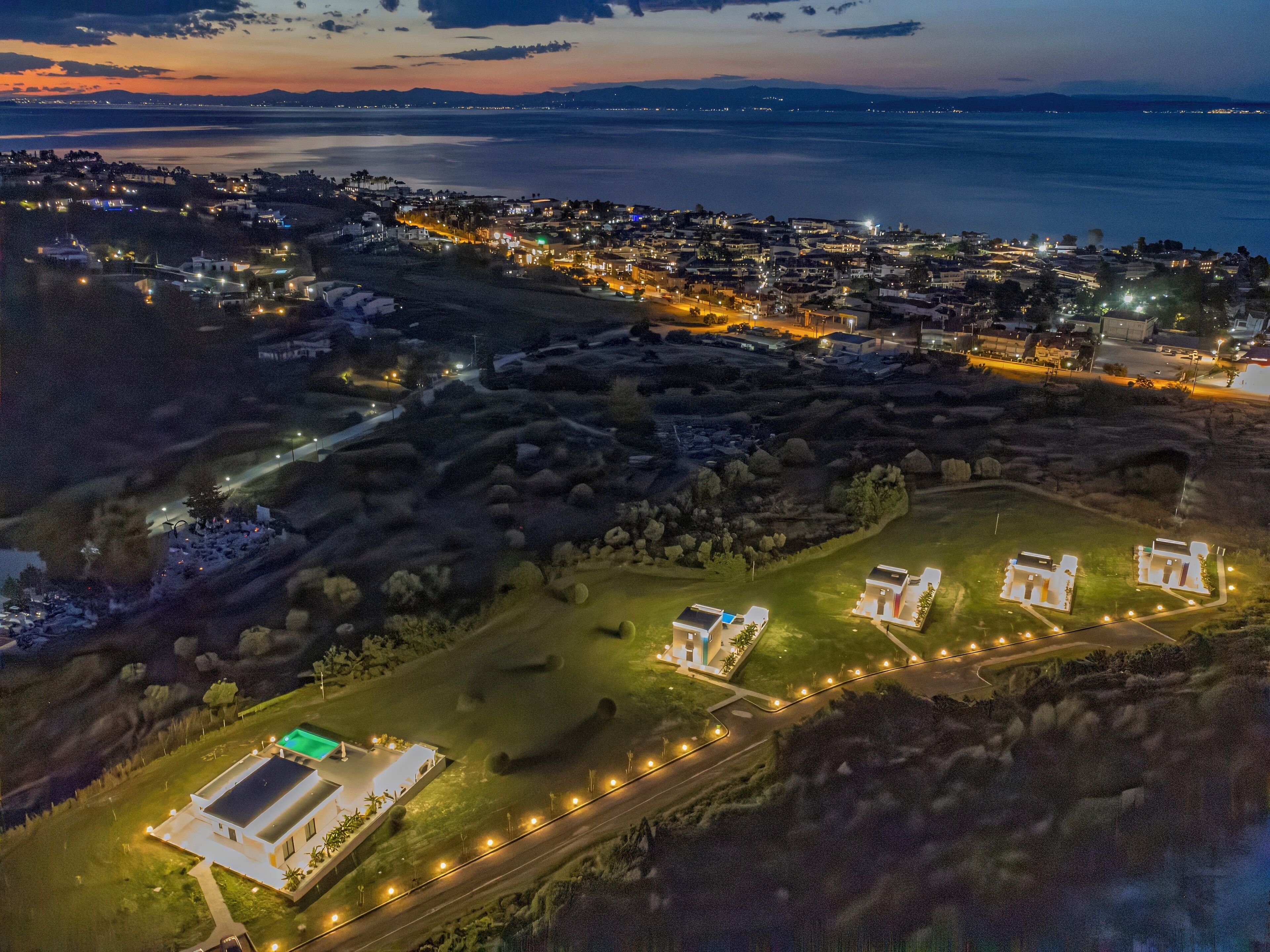 An aerial view of a coastal town at dusk with illuminated streets, a marina, and residential areas.