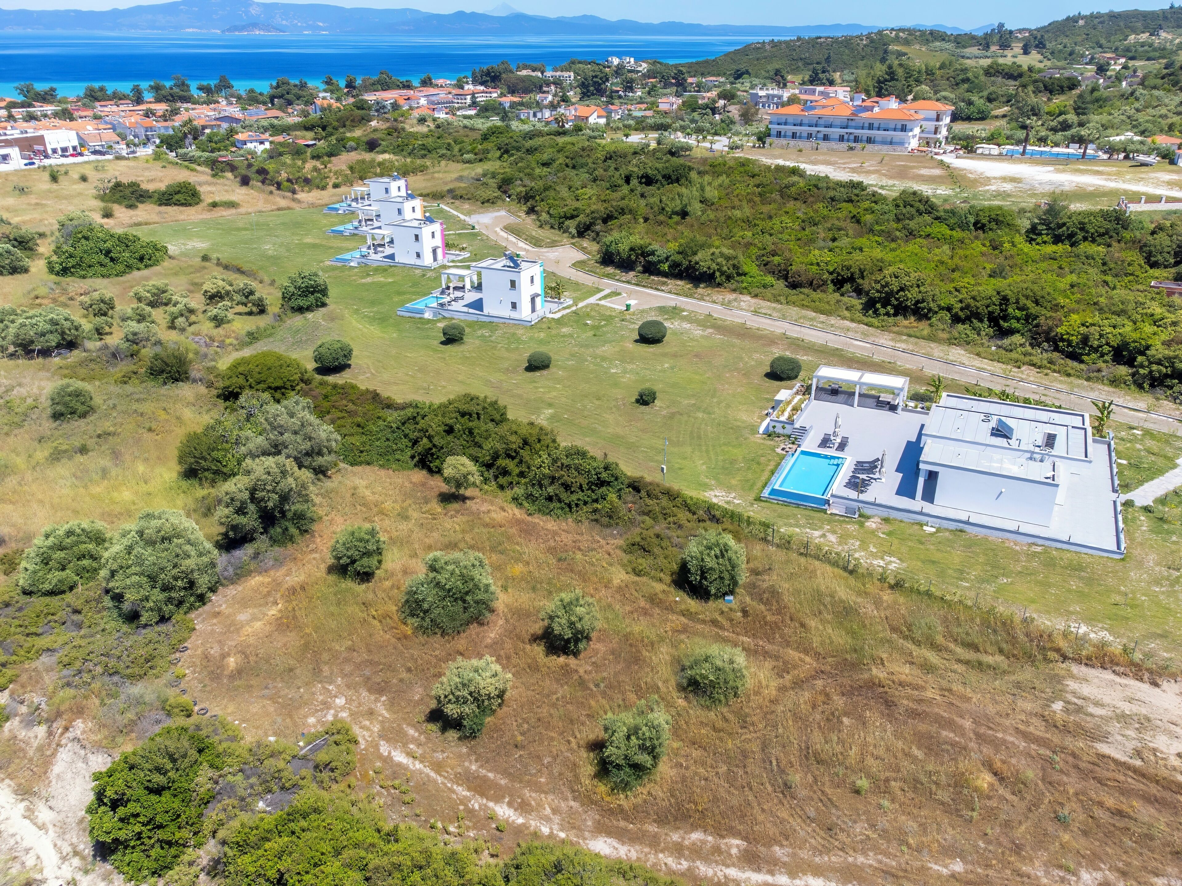 An aerial view of a residential area with modern houses, a swimming pool, and a hilly landscape with a view of the sea.