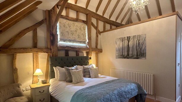 A bedroom with a sloped ceiling, exposed wooden beams, a bed with a blue blanket, a nightstand with a lamp, a chair, and a radiator. - The Red Cow Inn (Royston)